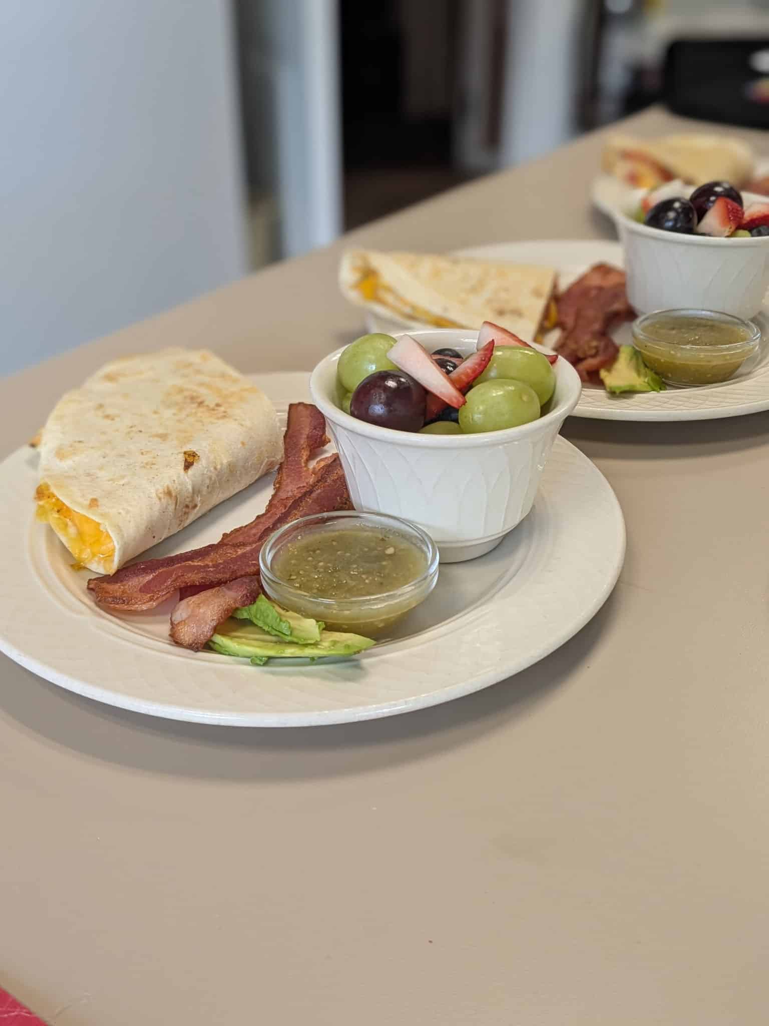 Brunch plate: wrap with cheese, crispy bacon, avocado slices, greens, and a small green salsa; fruit and olive cup on the side.
