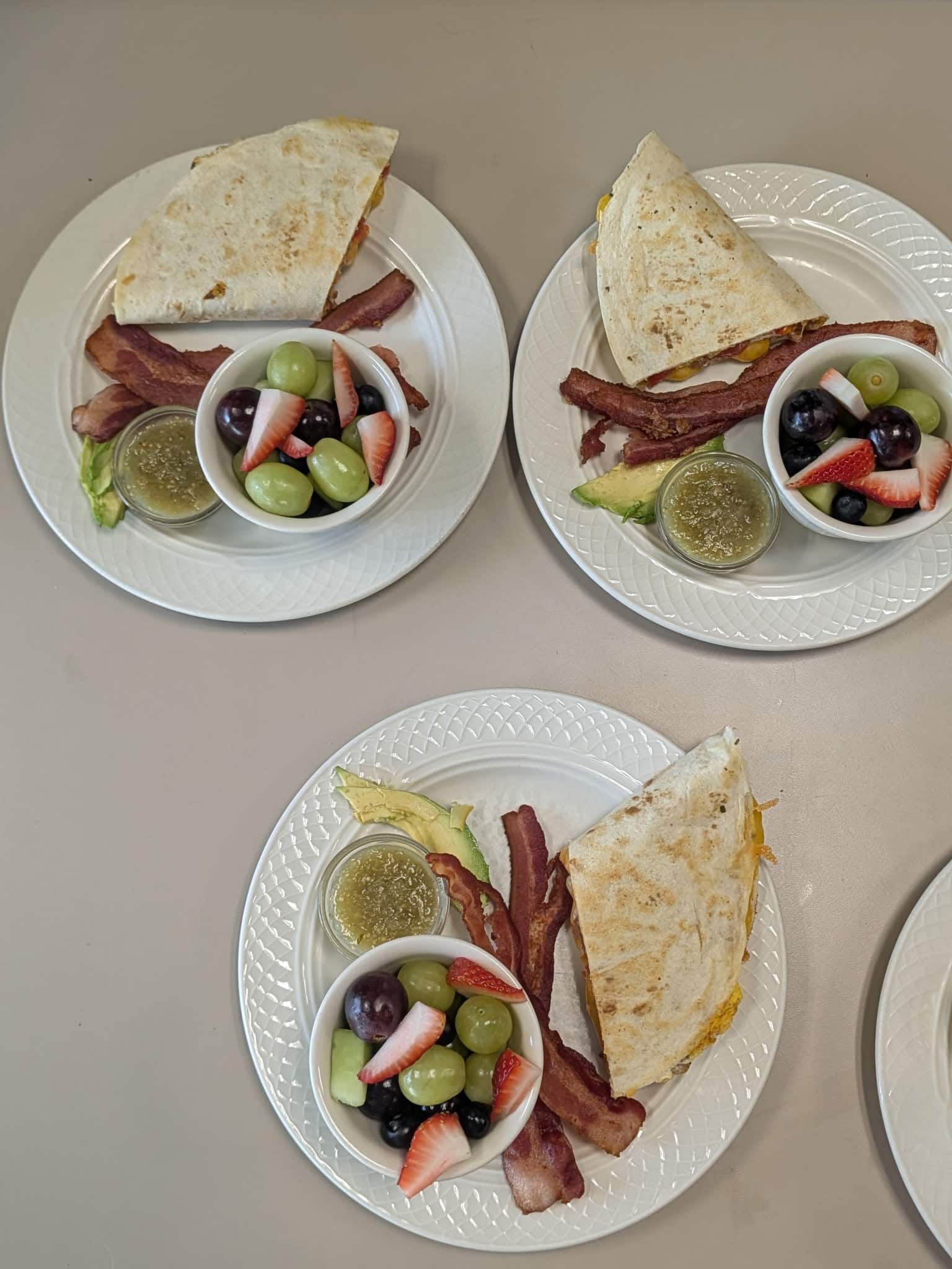 Three plates each feature a tortilla wrap with bacon, plus a small bowl of fruit (strawberries, grapes, blueberries) and a cup of green sauce on white plates.