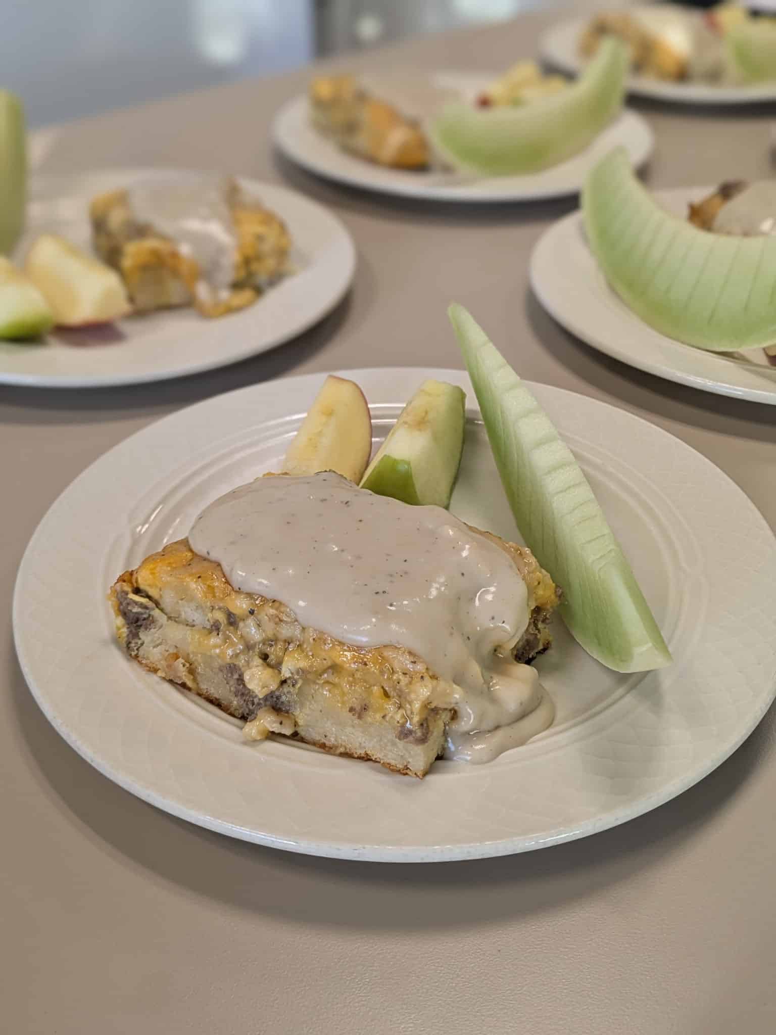 Slice of savory quiche with creamy mushroom sauce, served with apple wedges and a green melon wedge on a white plate.
