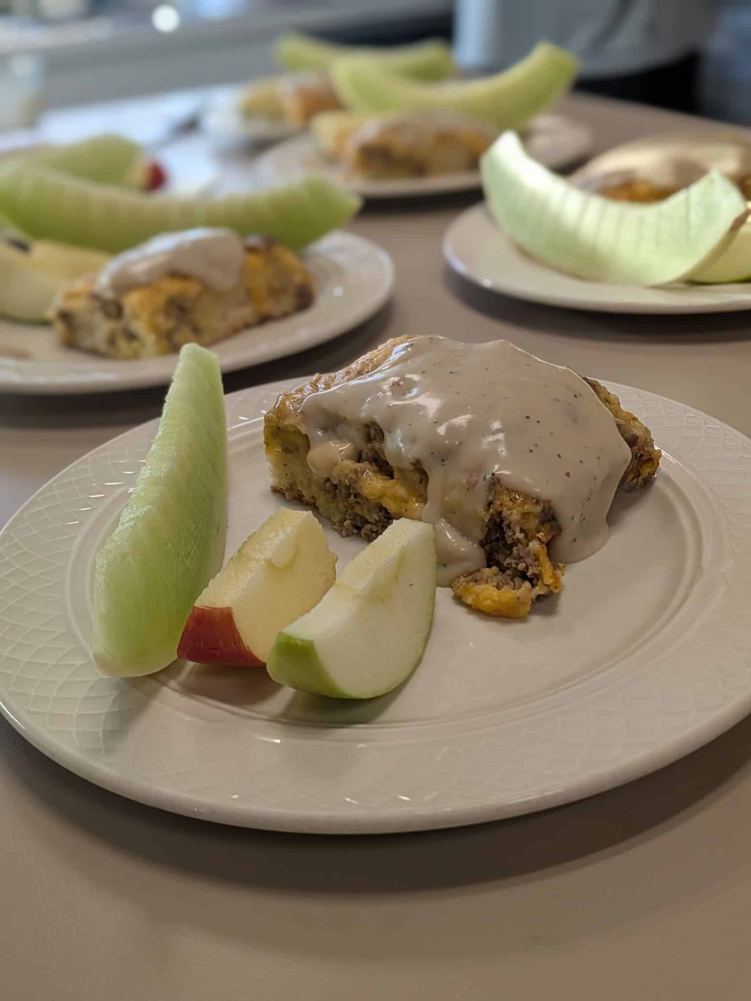 A plate with a square slice of casserole topped with beige gravy, surrounded by melon wedges and apple slices; blurred plates with more melon in the background.