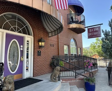 Brick guesthouse entrance with a purple door, arched window, a dog statue by the steps, and purple planters.