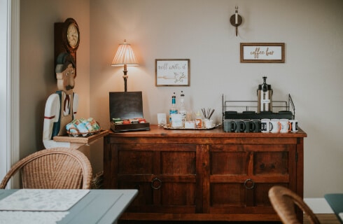 A room with a wood chest of drawers with a coffee and tea center on the top, tables and chairs, and a clock and lamps on the walls.