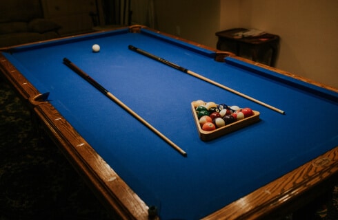 A billiard table with blue felt, a wooden frame, colorful billiard balls in a triangle rack, and two pool cues along the sides of the rack.