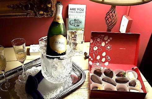 Two champagne glasses full of Champagne and chilled Champagne bottle on silver tray next to box of chocolates