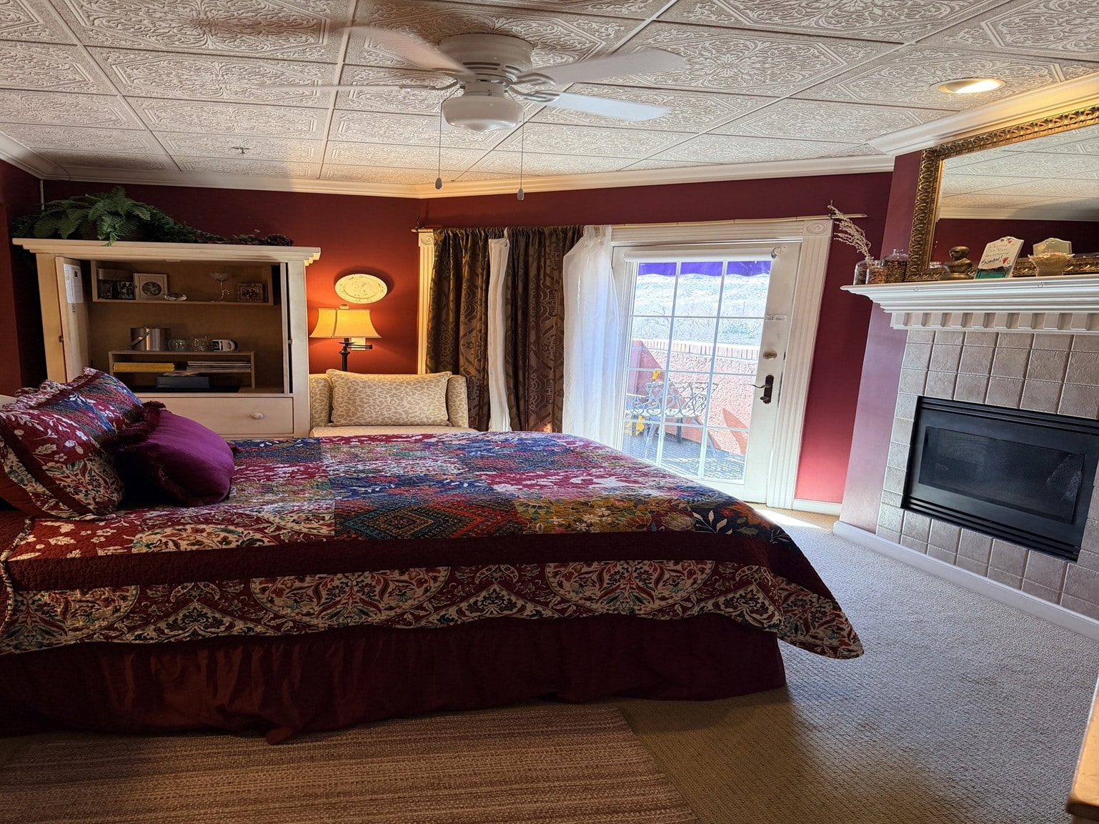 Cozy bedroom with a colorful quilted bed, burgundy walls, and sliding glass door to a patio beyond the bedframe