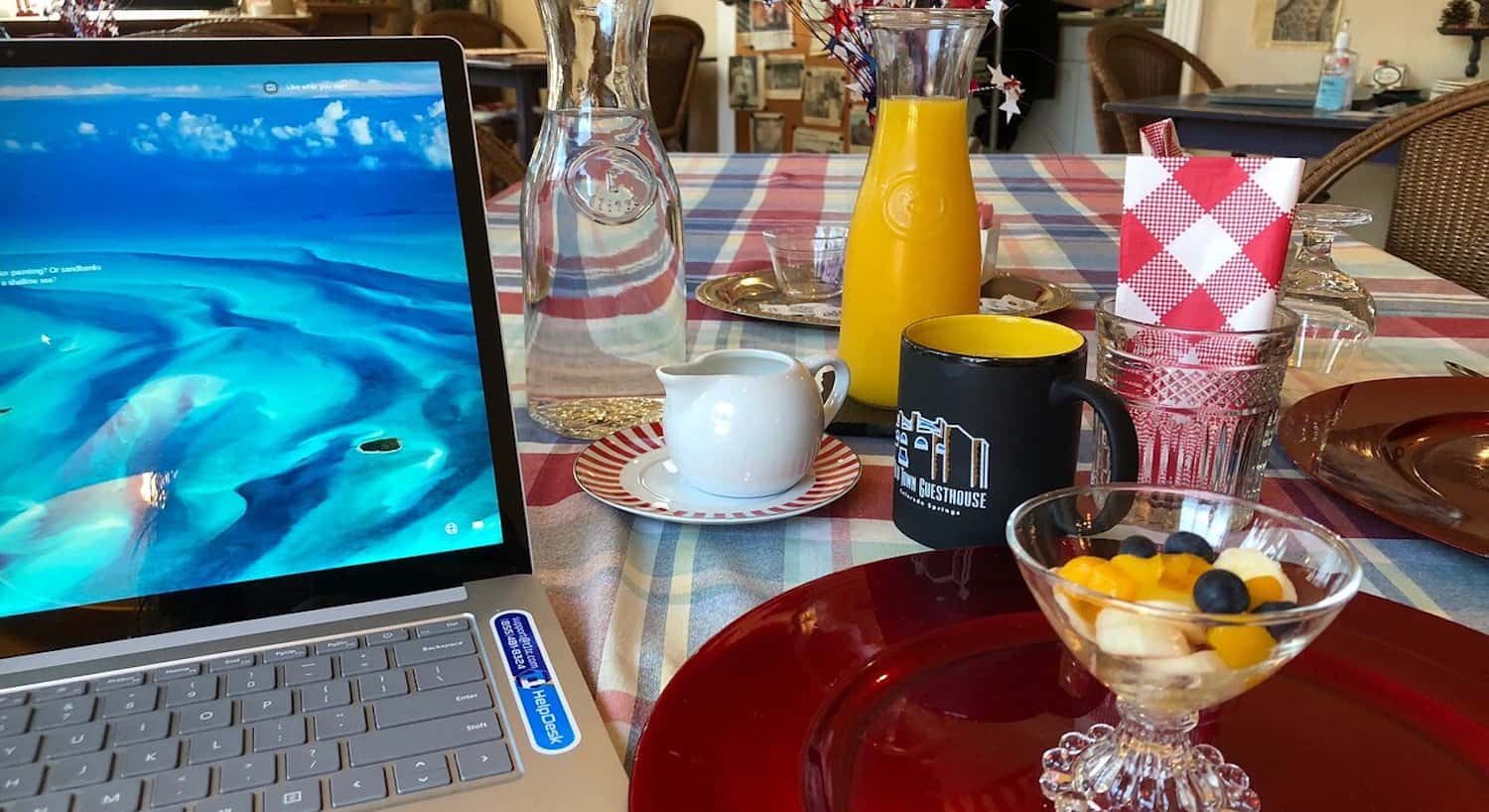 Close up view of a dining table with multicolored tablecloth, red plate with fruit cup, laptop, coffee cup, and carafes with water and orange juice