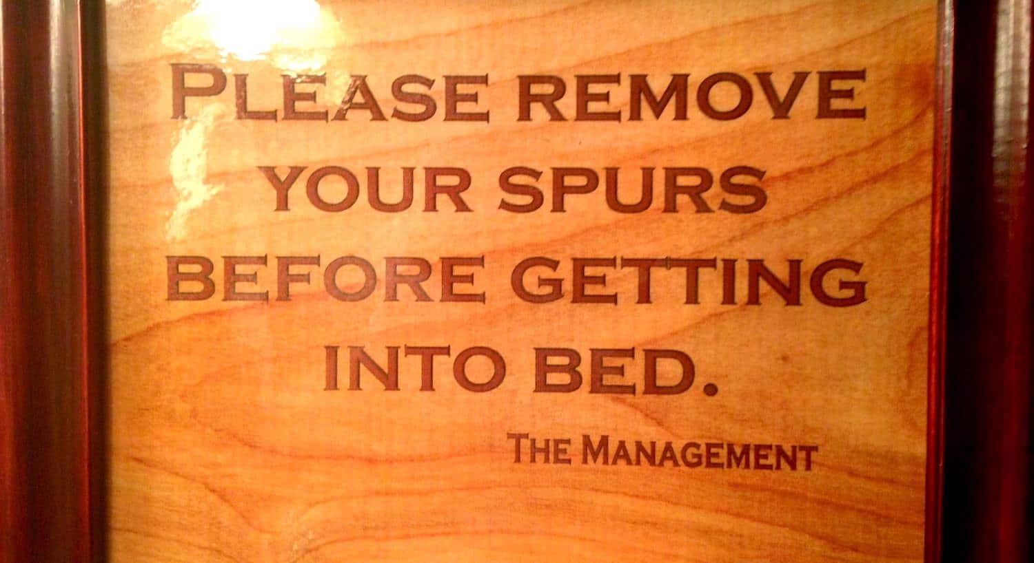 Close up view of wooden sign that reads Please remove your spurs before getting into Bed