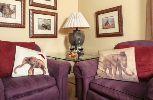 Bedroom with yellow walls, light colored carpeting, two purple upholstered armchairs, and glass end table with lamp