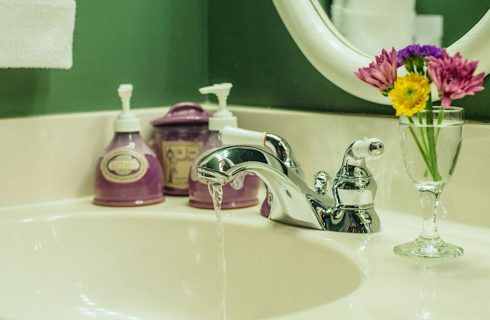 Close up view of running water out of chrome faucet with purple soap and lotion bottles and small glass vase with purple, pink, and yellow flowers