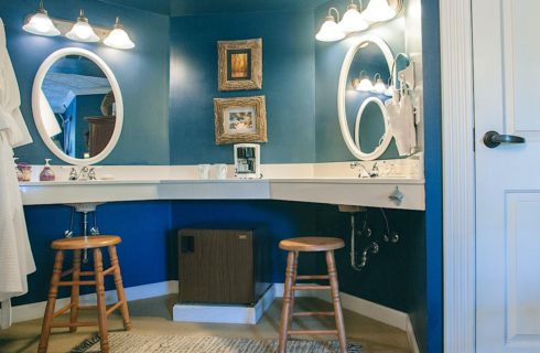 Bathroom with blue walls, double sinks, large counter, two oval mirrors, and a mini-fridge