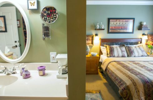 Bedroom with green walls, light colored carpeting, wood and wrought iron headboard, multicolored bedding, wooden nightstands, and view into bathroom with chrome faucet and oval mirror