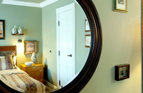 Close up view of large round mirror with wood and wrought iron headboard, multicolored bedding, and wooden nightstand in the reflection