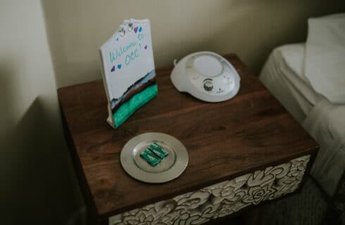 Close up view of a wooden carved nightstand next to a bed with a sign that says Welcome to OCC, a sound machine and a tray of mints on the table.