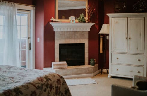 Bedroom with red walls, light colored carpeting, a bed with multicolored bedding, a tall white wardrobe on one wall, a fireplace with a white surround and mantle, and French doors opening to outside.