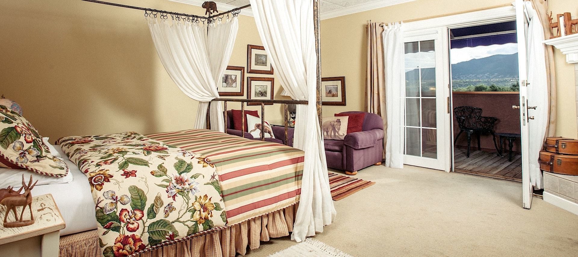 Bedroom suite with cream walls, light colored carpeting, four poster canopy bed with white curtains, purple upholstered armchairs, and opened door to balcony