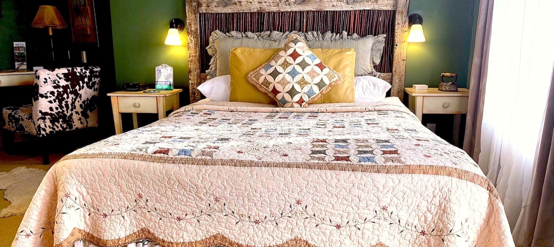 Bedroom with green walls, light colored carpeting, antique wooden headboard, multicolored bedding, and white wooden nightstands