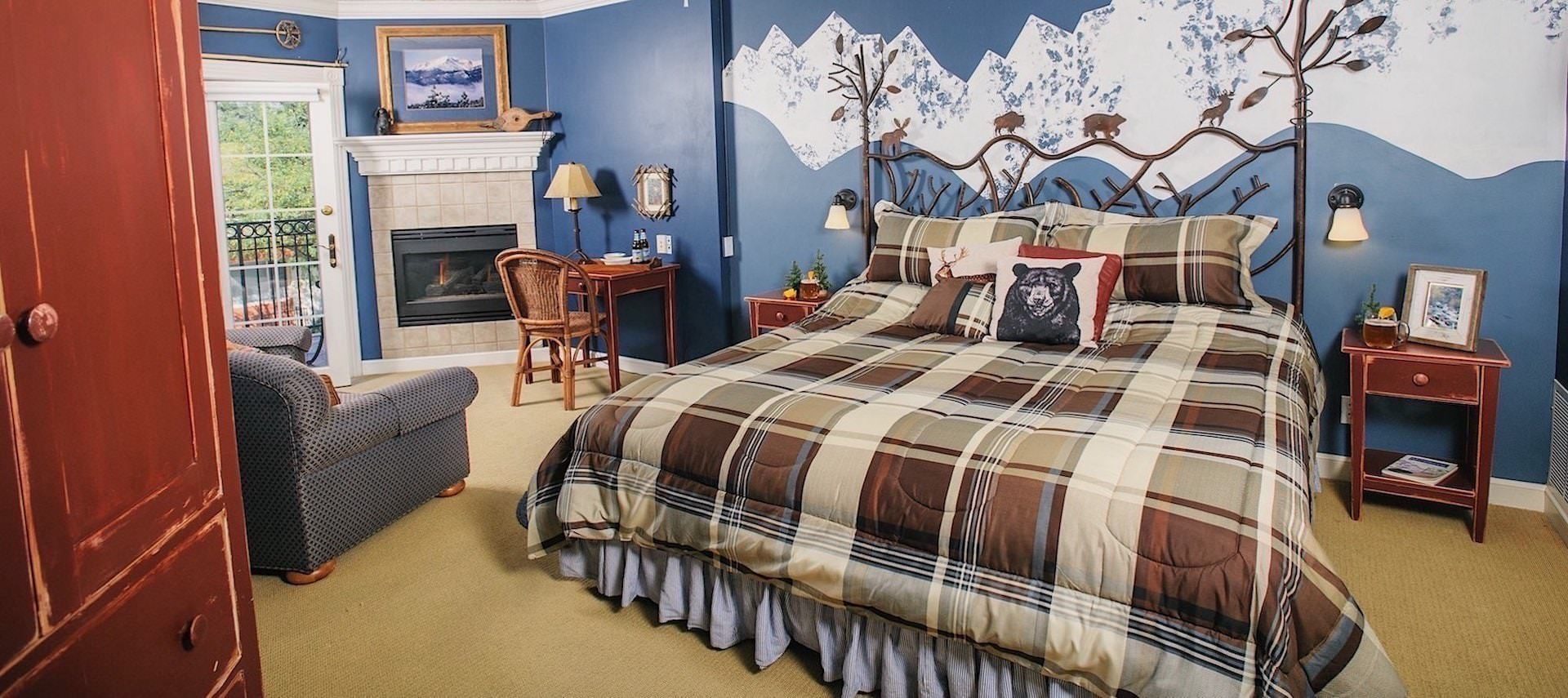 Bedroom with light colored carpeting, blue walls with white mountain range on ascent wall, decorative wrought iron headboard, multicolored plaid bedding, sitting area, desk, and fireplace