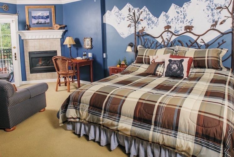 Bedroom with light colored carpeting, blue walls with white mountain range on ascent wall, decorative wrought iron headboard, multicolored plaid bedding, sitting area, desk, and fireplace
