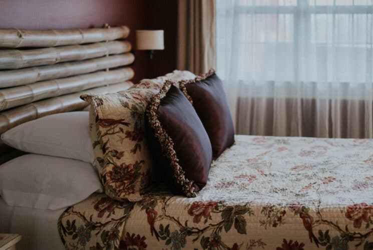 Bedroom with red walls, large bamboo rod headboard, multicolored bedding, and nightstands, and lamps on the walls on either side of the bed.