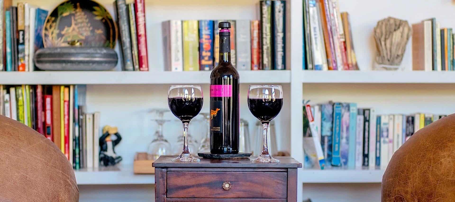 Close up view of two wine glasses full of red wine and bottle of wine on small wooden table with shelves of books in the background