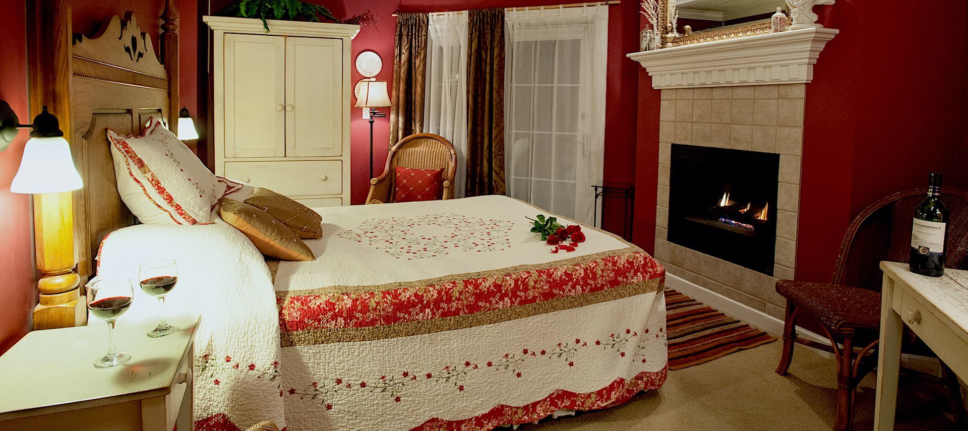 Bedroom with red walls, light colored carpeting, large wooden headboard, red and white bedding, white wooden nightstand and armoire, and fireplace