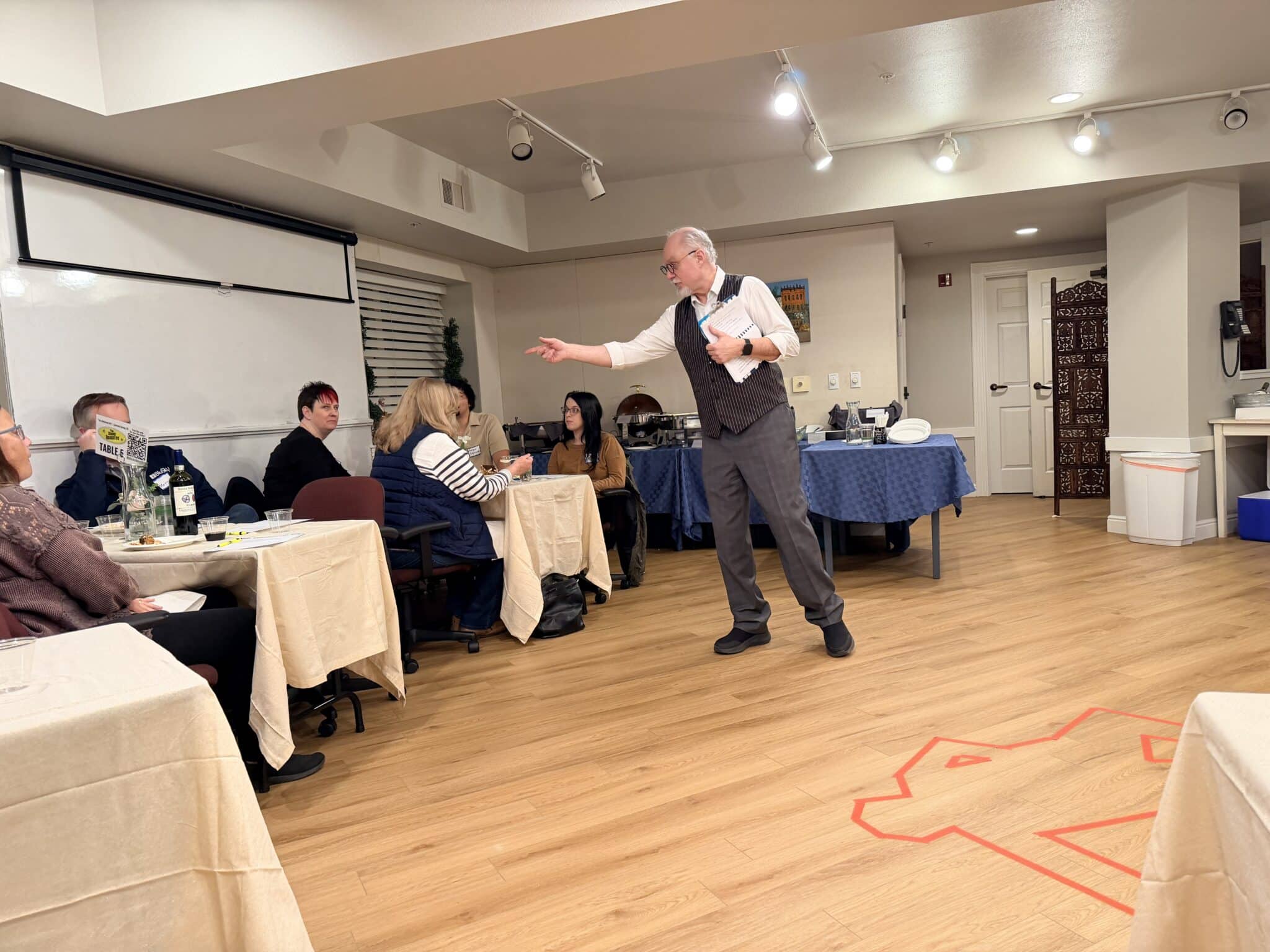 A man in a vest and tie speaks to a seated group at round tables in a banquet room during a social event.
