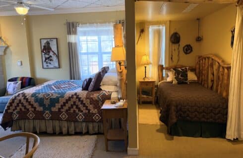 Split-bedroom room with a queen bed on the left featuring a Southwestern-style quilt and a rustic log-frame bed on the right, divided by a partial wall and warm lamps.
