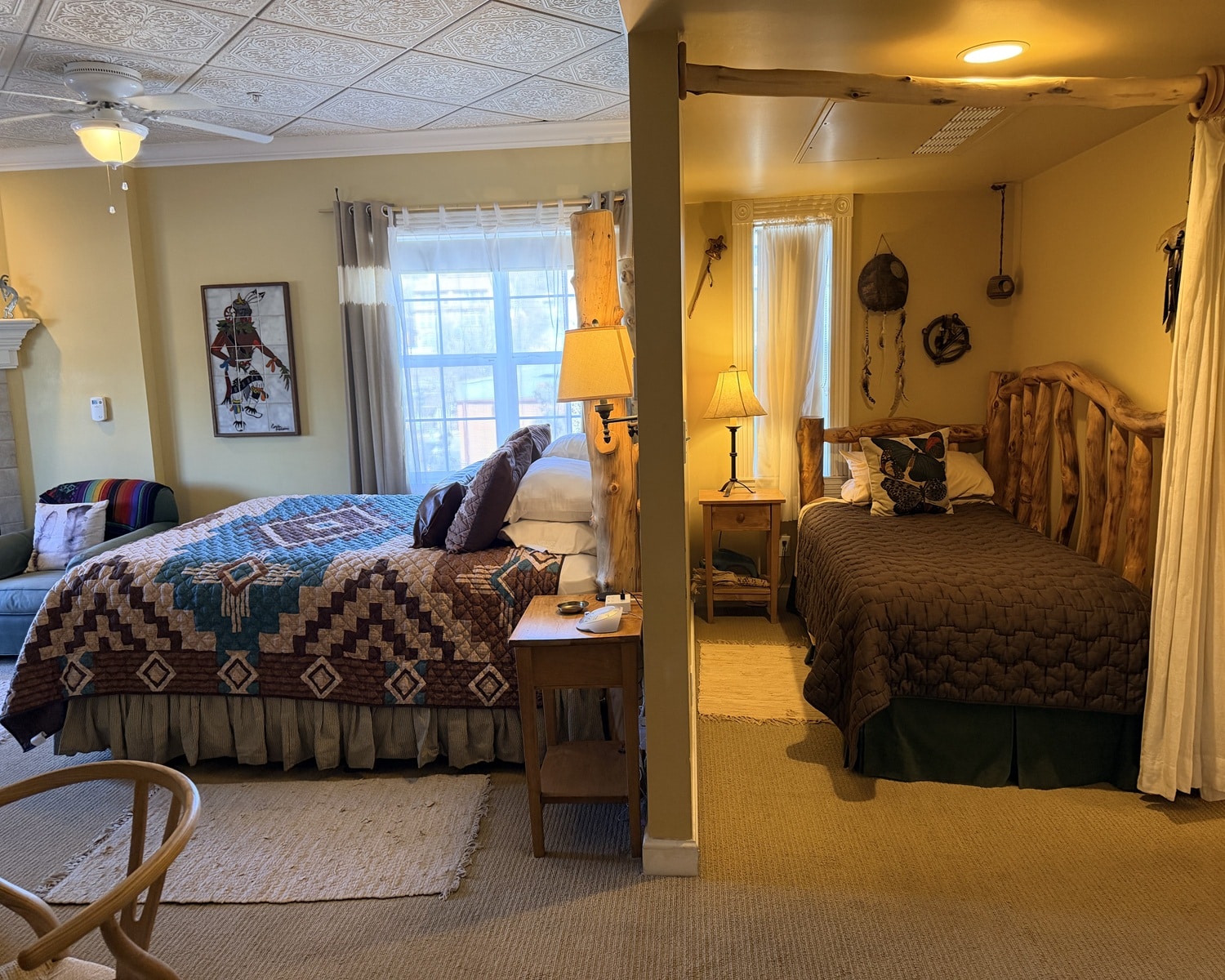 Split-bedroom room with a queen bed on the left featuring a Southwestern-style quilt and a rustic log-frame bed on the right, divided by a partial wall and warm lamps.