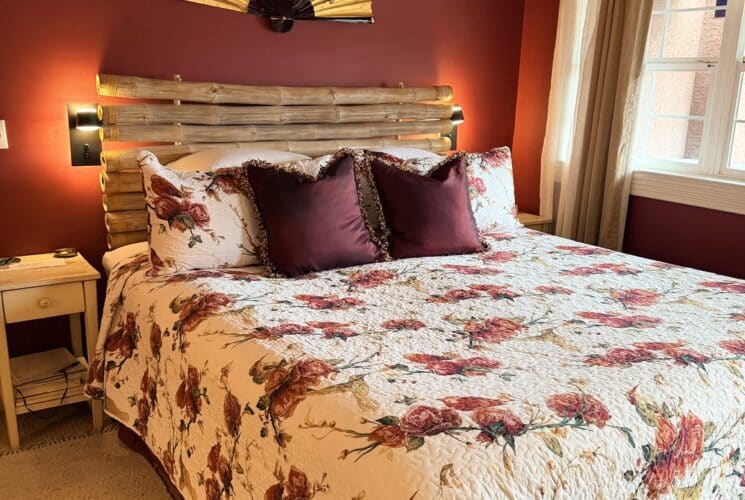 Cozy bedroom with burgundy walls, a bamboo headboard, floral quilt, and plum throw pillows; bedside tables and soft window light.