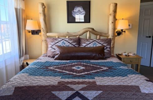 Rustic log bed with a geometric quilt, two bedside lamps, and a framed hat artwork above on a yellow wall.