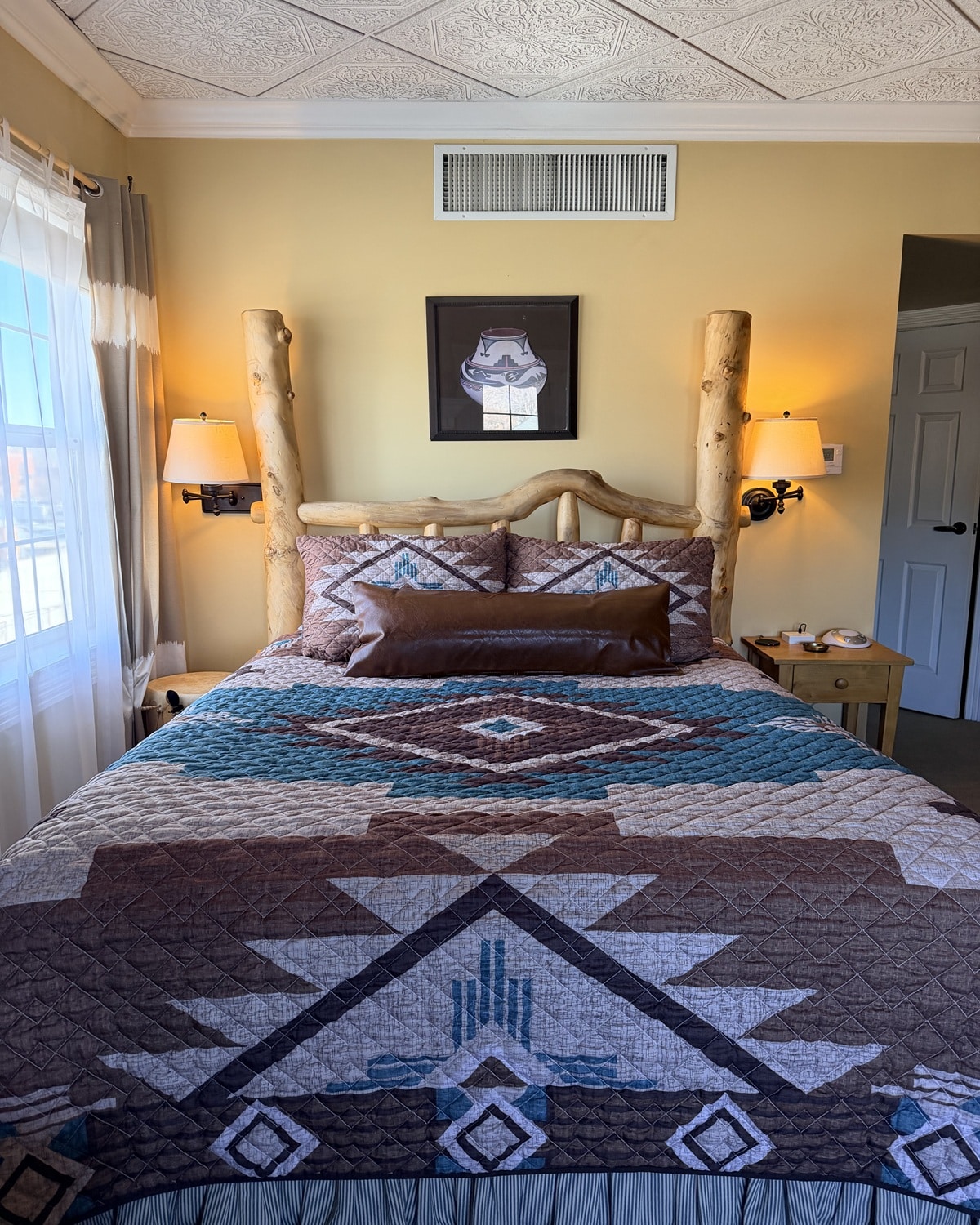 Rustic log bed with a geometric quilt, two bedside lamps, and a framed hat artwork above on a yellow wall.