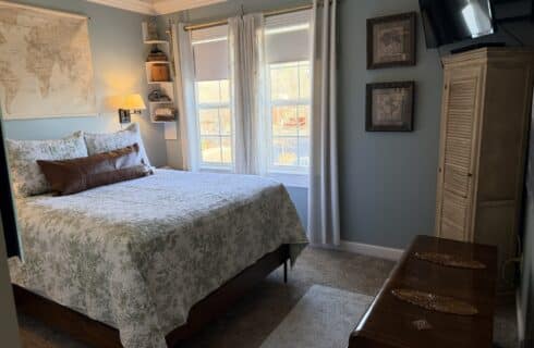 Cozy bedroom with a queen bed in floral green bedding, a brown bolster pillow, blue walls, and a world map on the wall above the bed; window with white curtains to the right and a small wall shelf with decor.