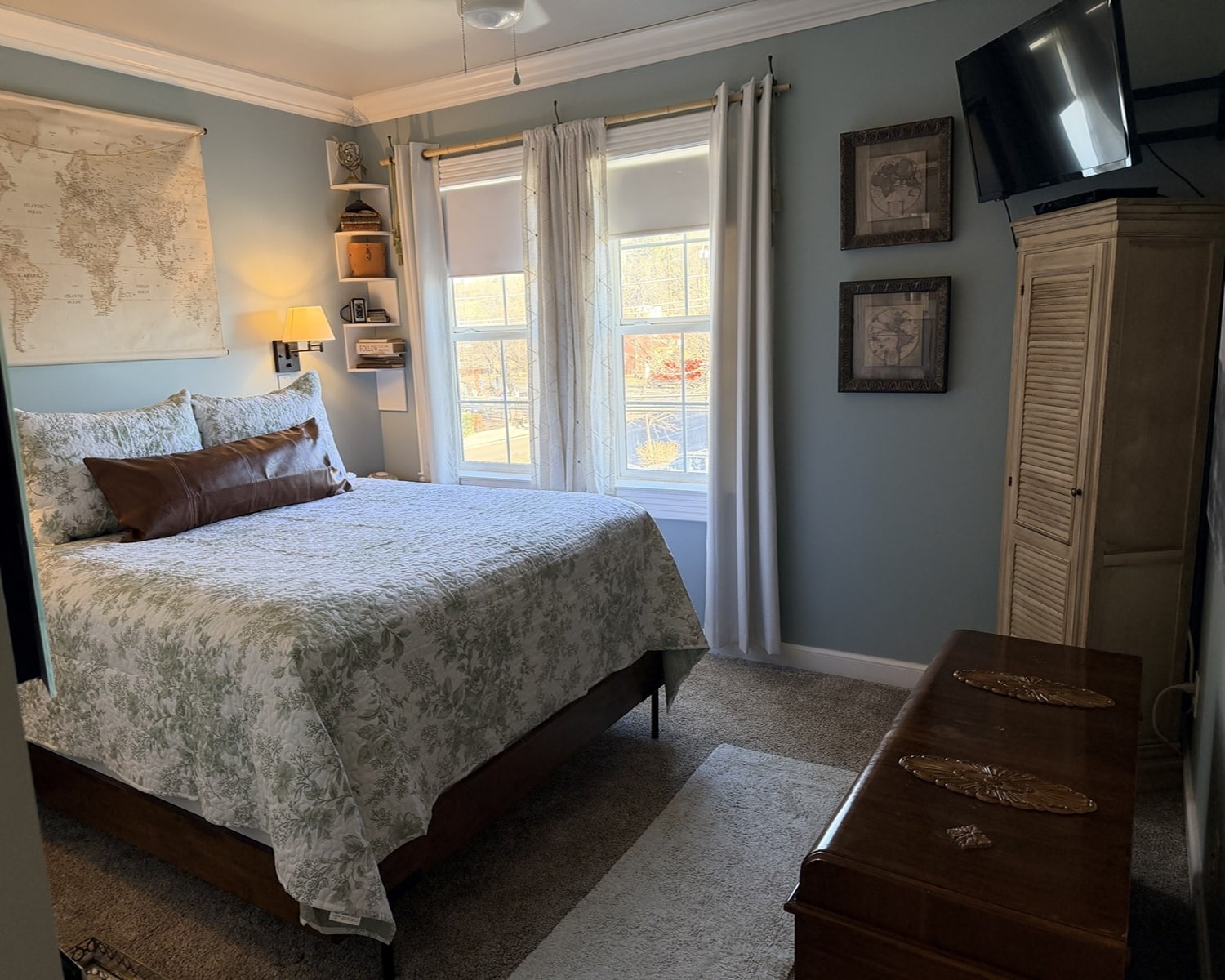Cozy bedroom with a queen bed in floral green bedding, a brown bolster pillow, blue walls, and a world map on the wall above the bed; window with white curtains to the right and a small wall shelf with decor.