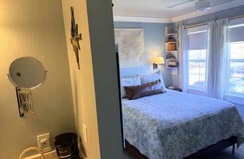 Cozy bedroom with blue walls, a queen bed with a light floral quilt, and a large window with white curtains. Shelf decor and a ceiling fan visible on the ceiling corner.
