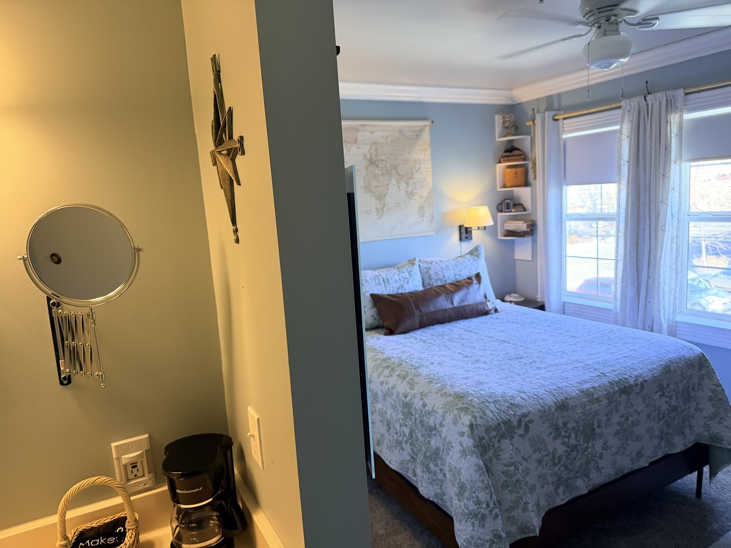 Cozy bedroom with blue walls, a queen bed with a light floral quilt, and a large window with white curtains. Shelf decor and a ceiling fan visible on the ceiling corner.