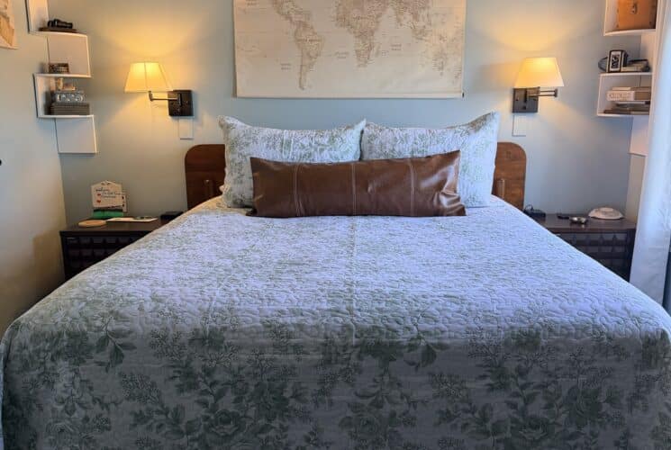 Queen bed with pale blue floral quilt and brown lumbar pillow under a world map wall art.
