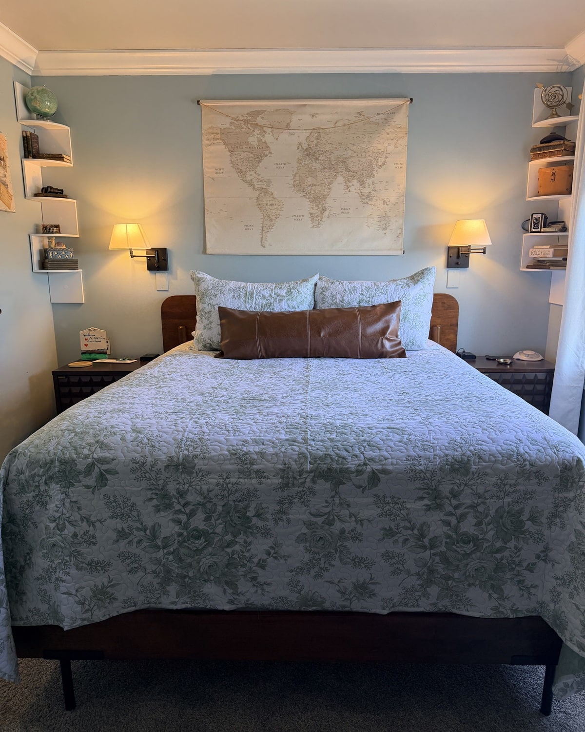 Queen bed with pale blue floral quilt and brown lumbar pillow under a world map wall art.
