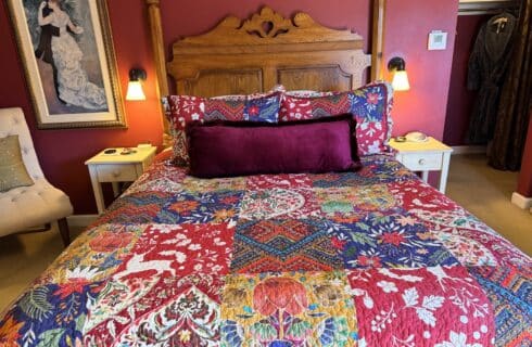 Bed with colorful floral quilt and burgundy velvet bolster pillow against a carved wooden headboard. Two white nightstands with lamps flank the bed on red walls.