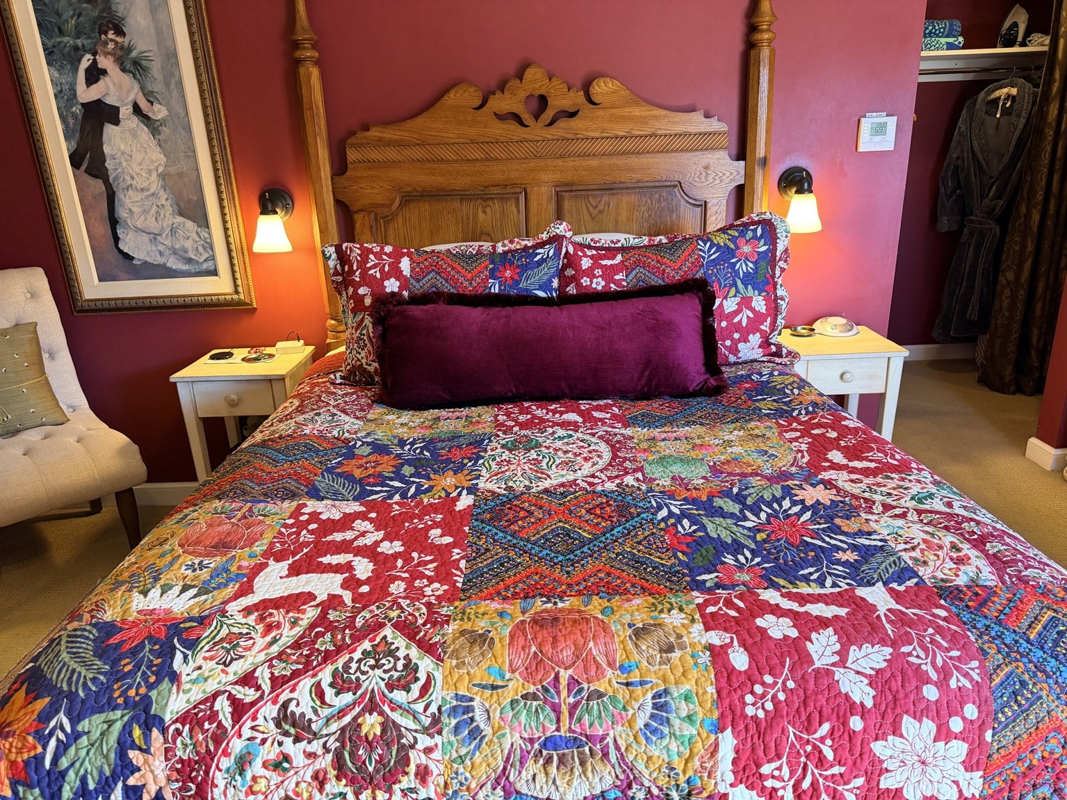 Bed with colorful floral quilt and burgundy velvet bolster pillow against a carved wooden headboard. Two white nightstands with lamps flank the bed on red walls.