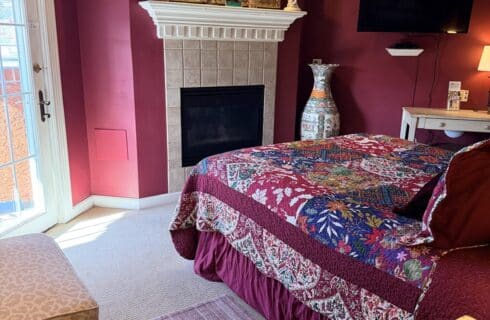Hotel bedroom with magenta walls, a white fireplace, and an ornate gold-framed mirror above it; a colorful quilted bed to the right.