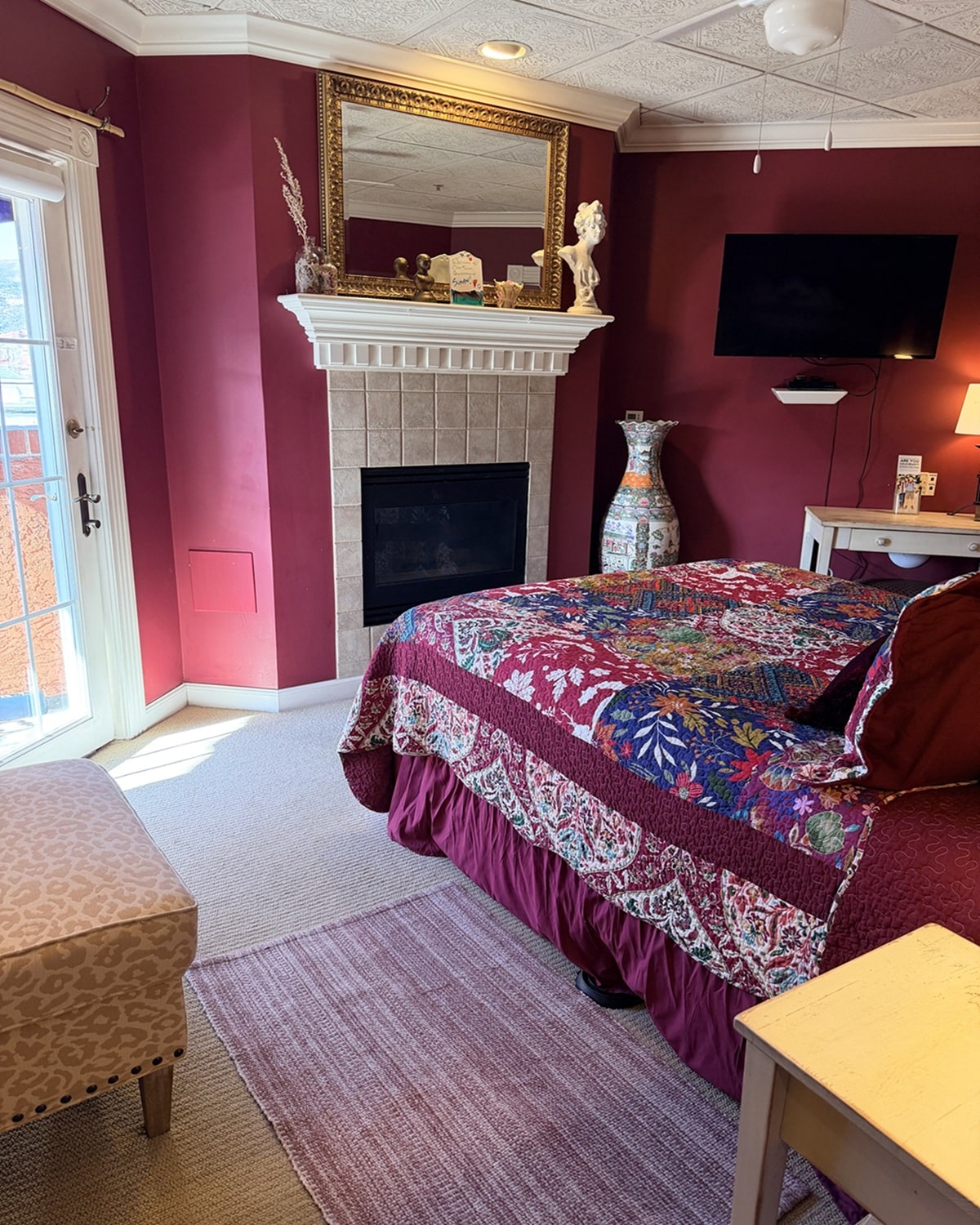Hotel bedroom with magenta walls, a white fireplace, and an ornate gold-framed mirror above it; a colorful quilted bed to the right.
